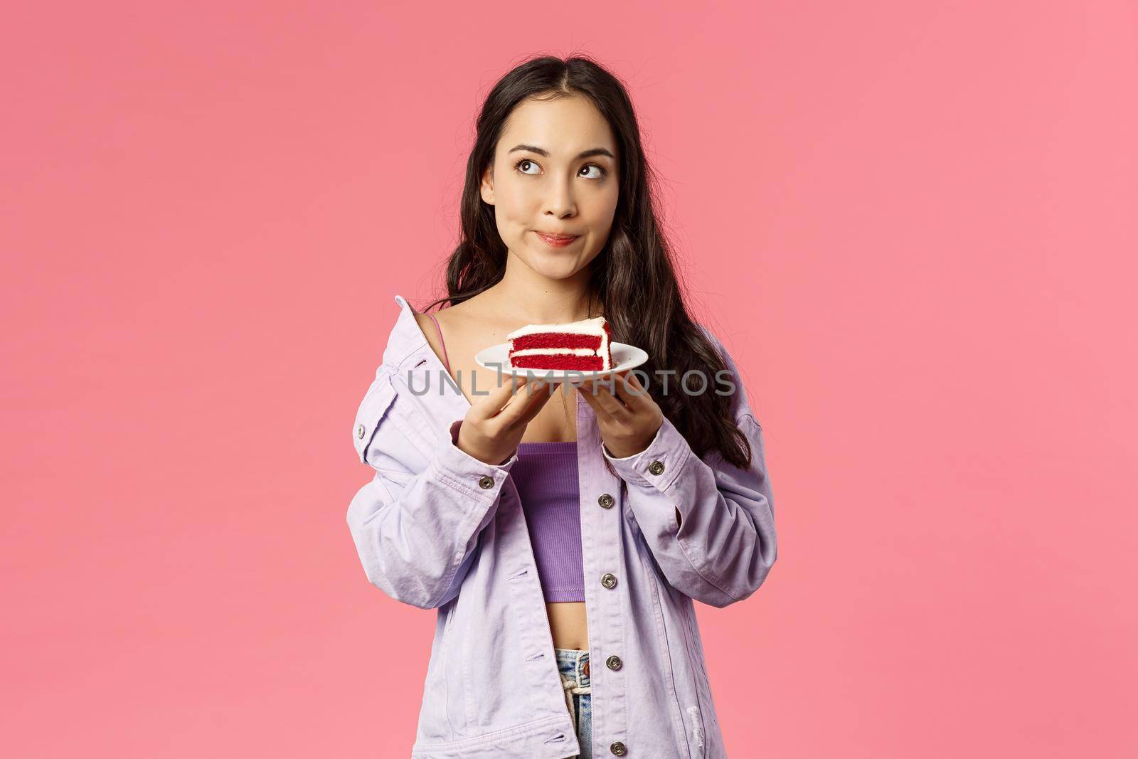 Troubled young asian girl wants to eat delicious piece cake but having doubts, hesitating of lots calories, smirk concerned look upper left corner thinking, making decision, pink background by Benzoix