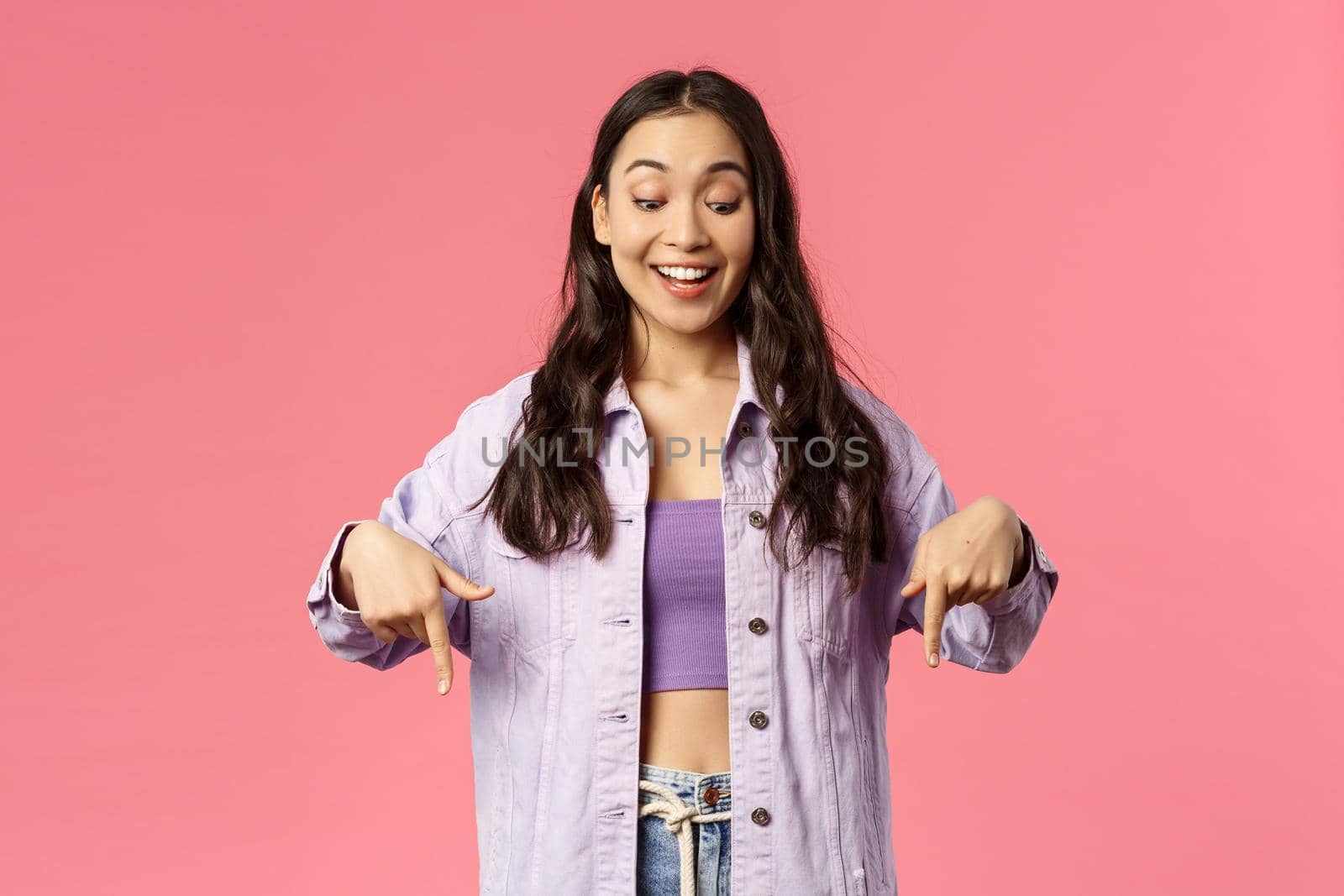 Portrait of curious good-looking, stylish female in denim jacket over crop-top, smiling excited and looking pointing fingers down at something interesting and unsual, stand pink background.