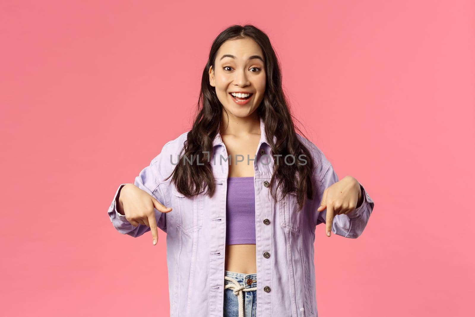 Portrait of excited, happy smiling korean girl in denim jacket, talking about cool new product in store, online shopping site or application, recommend click link, pointing fingers down.