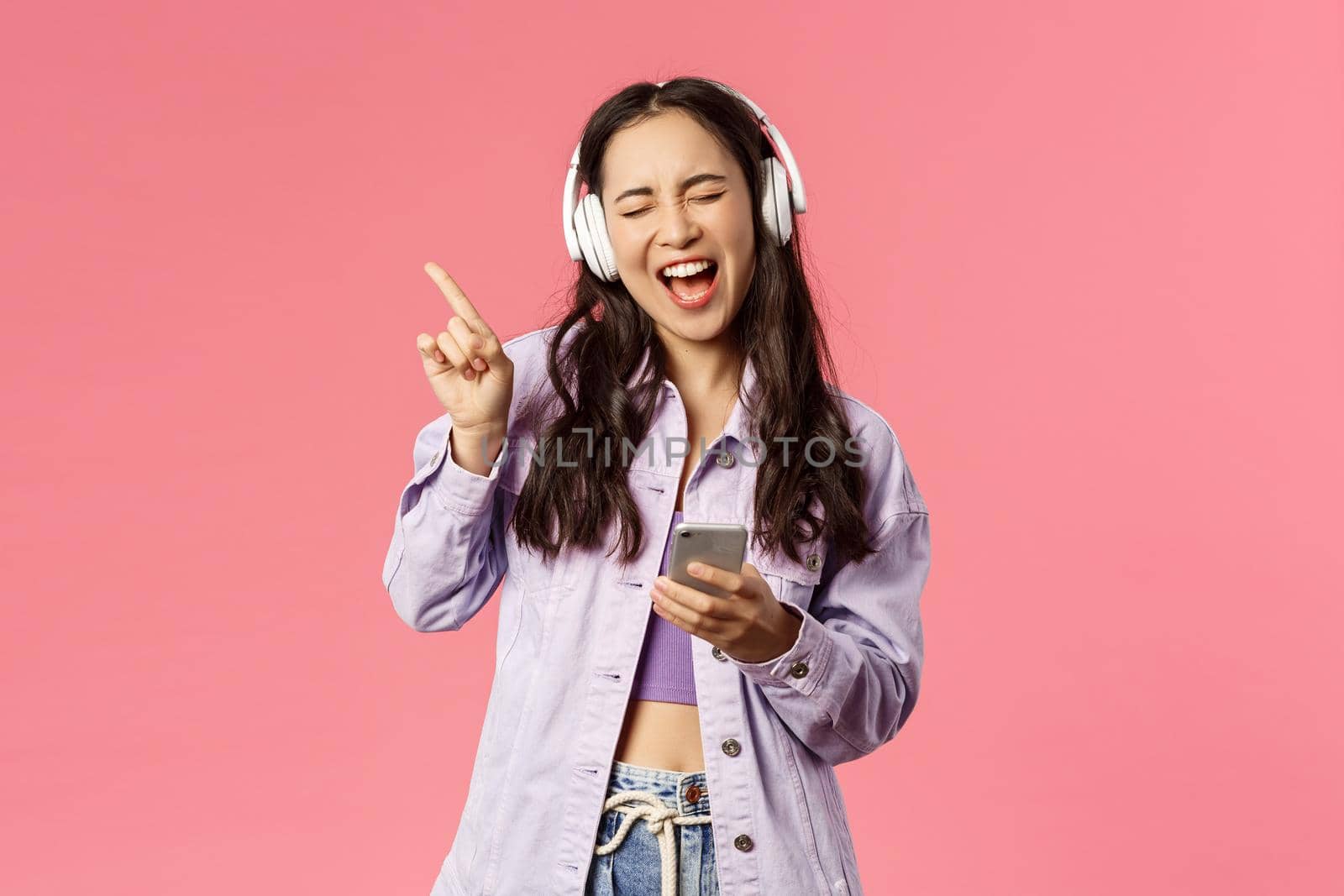 Waist-up portrait of excited and carefree stylish woman enjoying favourite songs, listening music in headphones, close eyes dancing to rhythm and singing along, hold smartphone by Benzoix