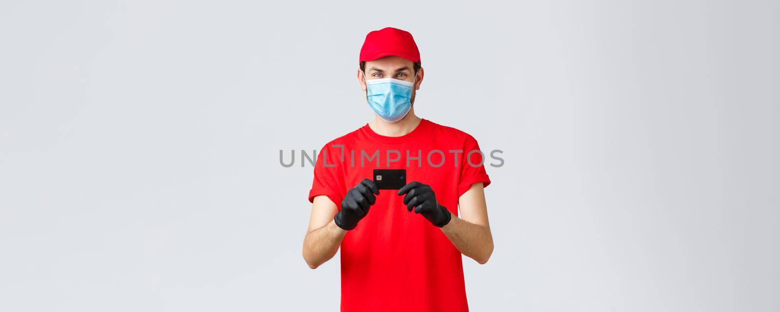 Contactless delivery, payment and online shopping during covid-19, self-quarantine. Cheerful courier in red uniform cap and t-shirt, wear medical face mask, gloves showing credit card, order food.