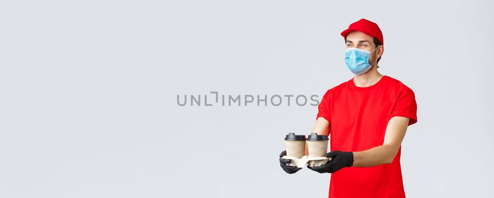 Takeaway, food and groceries delivery, covid-19 contactless orders concept. Pleasant courier in red uniform, face mask and gloves, giving out coffee to customer, stand grey background by Benzoix
