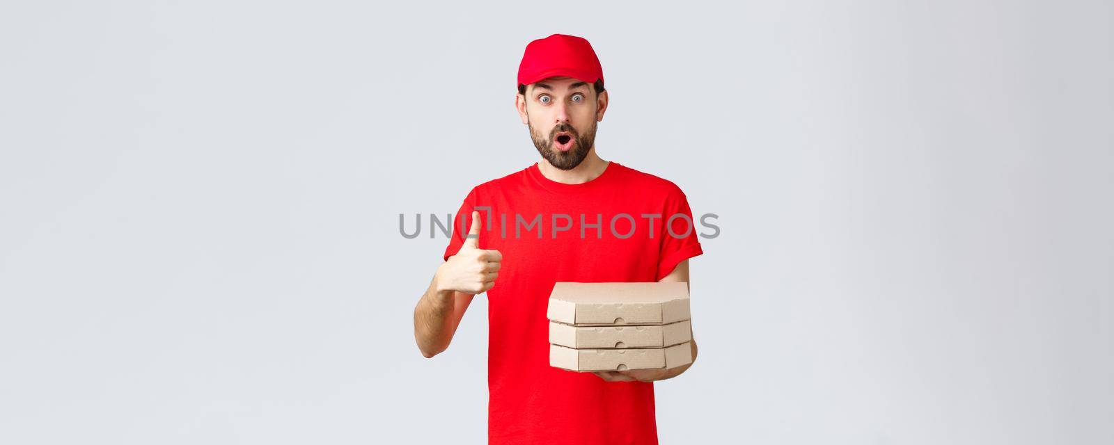 Food delivery, quarantine, stay home and order online concept. Surprised and amazed bearded courier in red t-shirt, cap gasping impressed, show thumb-up, bring order pizza, grey background.