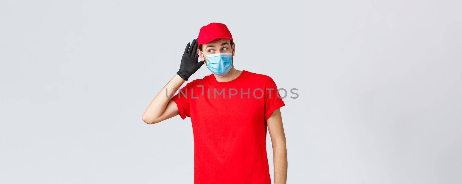 Groceries and packages delivery, covid-19, quarantine and shopping concept. Intrigued courier in red uniform, face mask and gloves, overhearing conversation, eavesdrop, hold hand near ear by Benzoix