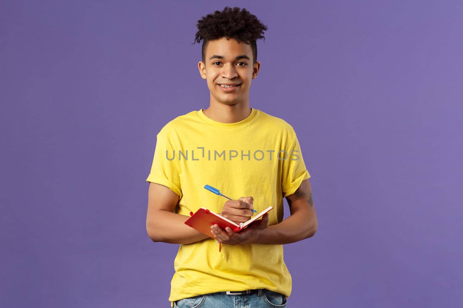 Portrait of young hispanic male student studying online courses, writing down lecture, making personal schedule or taking notes in planner, look camera enthusiastic and interested, purple background