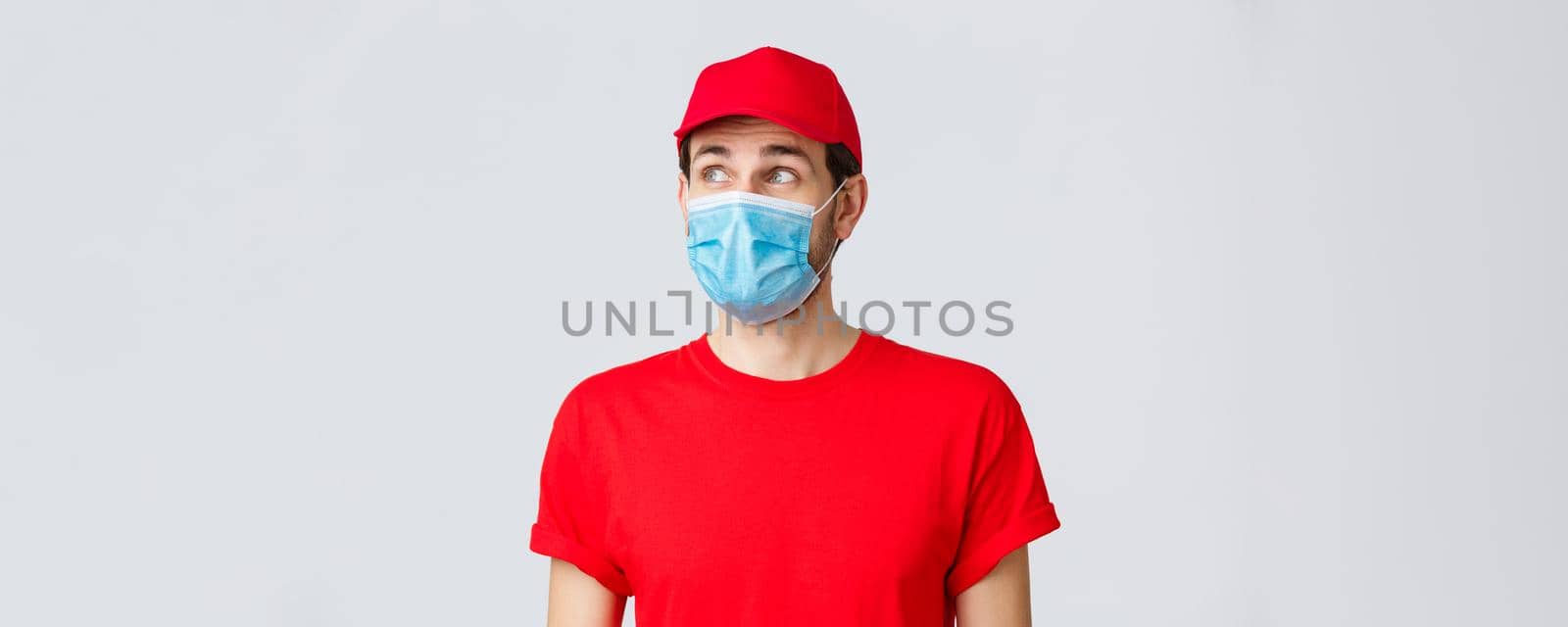 Contactless delivering, covid-19 and shopping concept. Thoughtful courier in red uniform cap, t-shirt and medical face mask with gloves, looking upper left corner, reading banner or advertisement by Benzoix