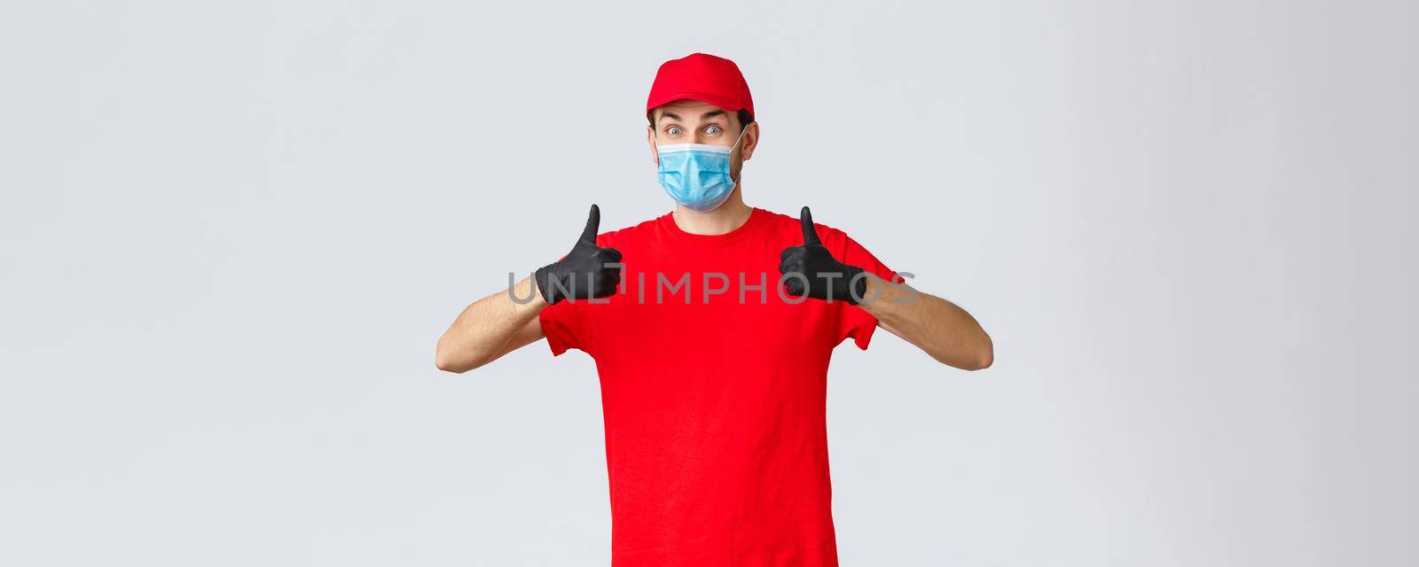 Covid-19, self-quarantine, shopping and shipping concept. Enthusiastic and happy courier in red uniform, gloves and face mask, support idea, thumb-up, recommend client contactless package delivery.