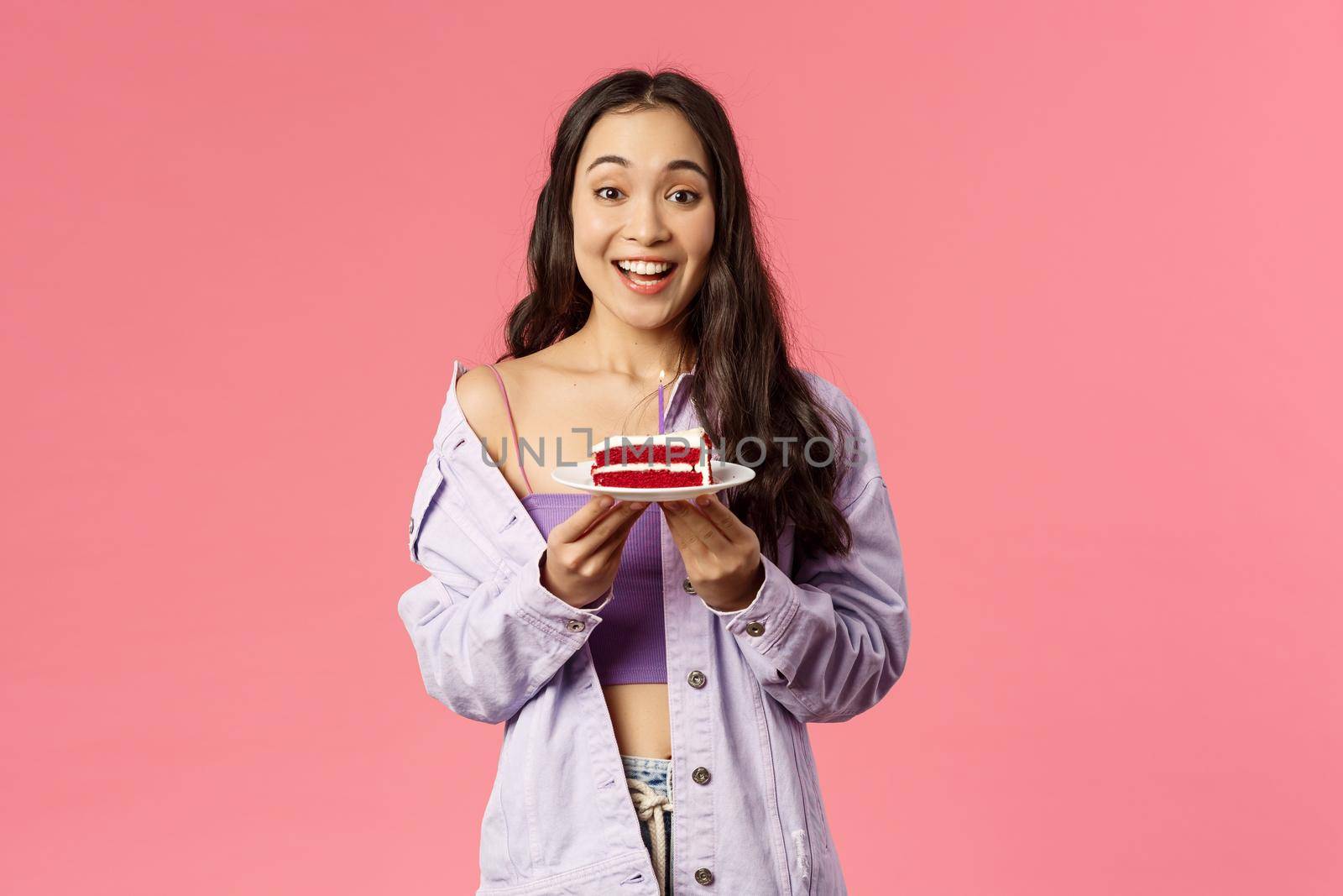 Portrait of cheerful, happy young cute woman baked a dessert special for you, holding piece of cake on plate, suggesting try it, smiling amused and excited, eating favorite food, pink background.