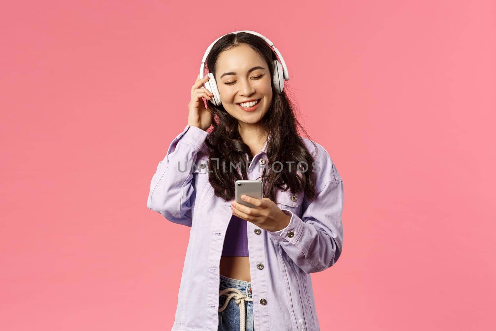 Portrait of stylish attractive and happy, smiling asian girl enjoying listening favourite songs in headphones, close eyes and laughing, found music station or funny podcast, hold mobile phone.