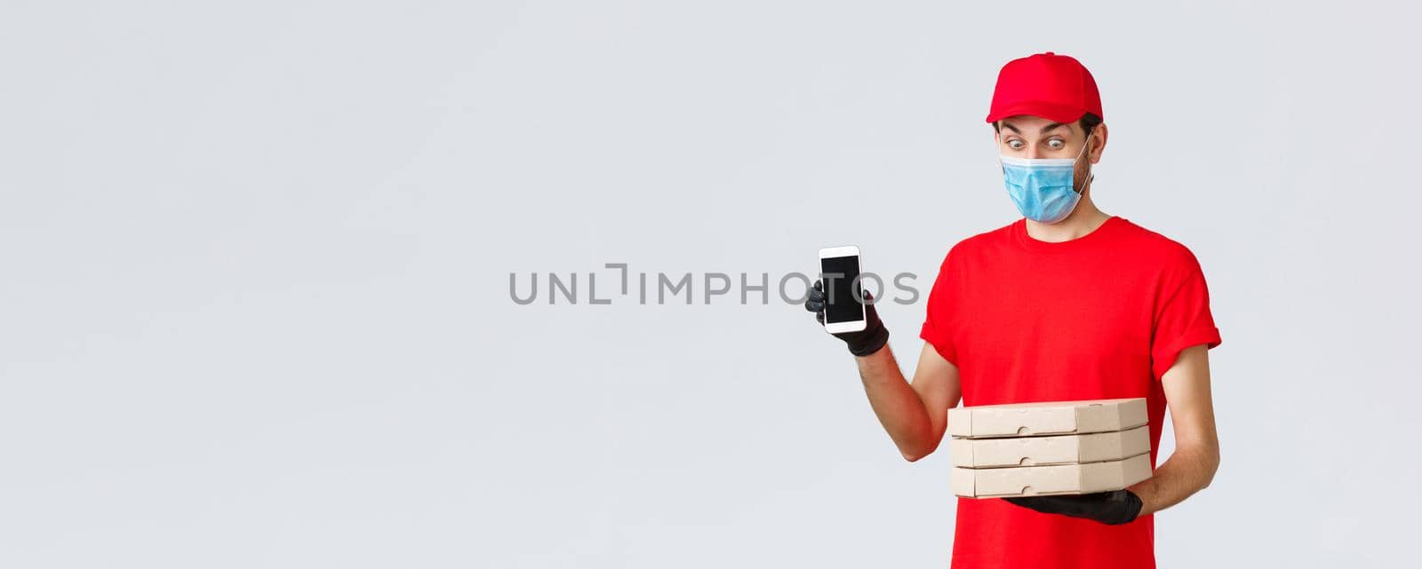 Food delivery, application, online grocery, contactless shopping and covid-19 concept. Amused funny courier in red uniform, face mask and gloves, showing smartphone screen app and hold pizza boxes by Benzoix