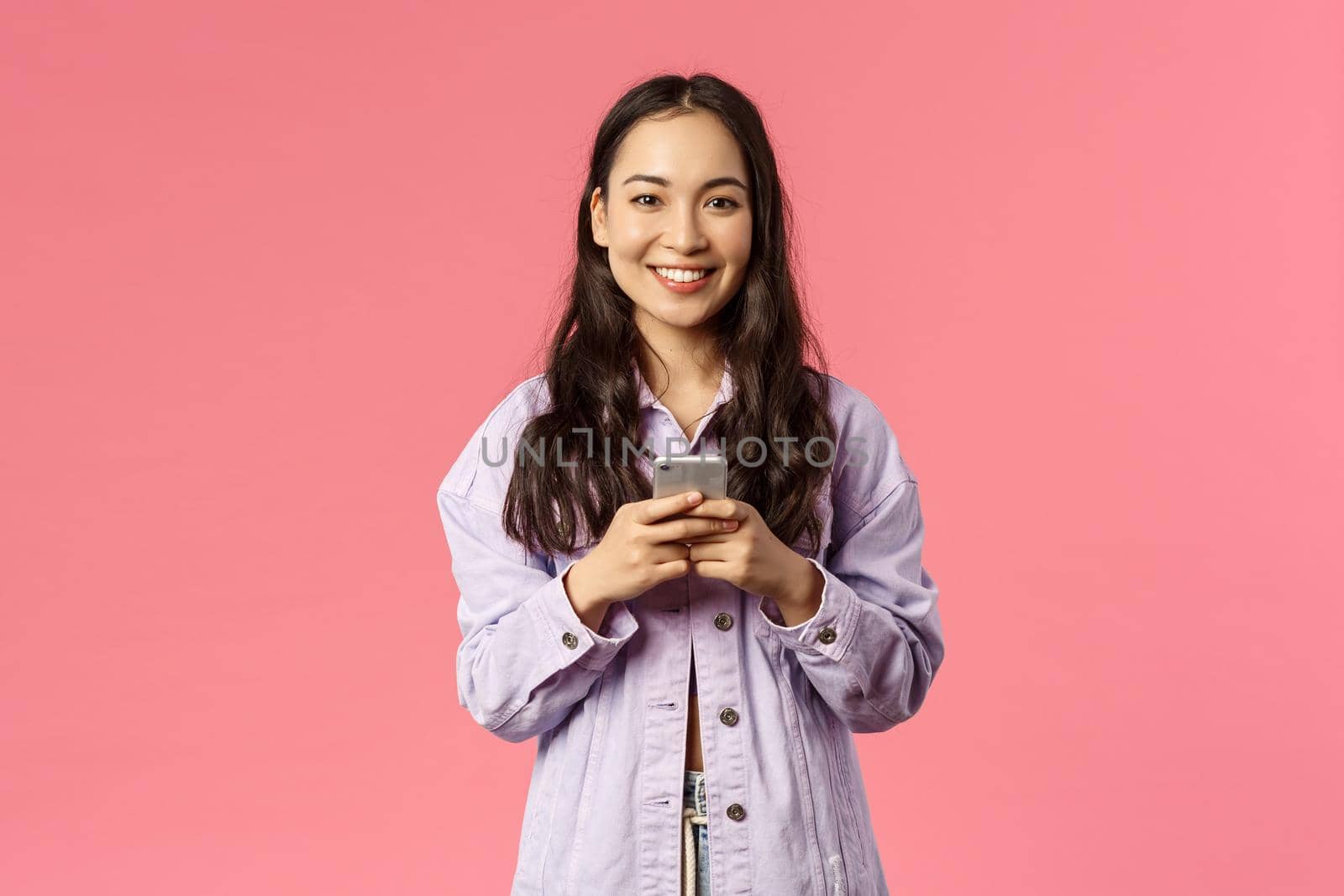 Online lifestyle, people and beauty concept. Cheerful stylish young girl using mobile phone and smiling pleased at camera, chatting during lockdown, use education app, pink background by Benzoix