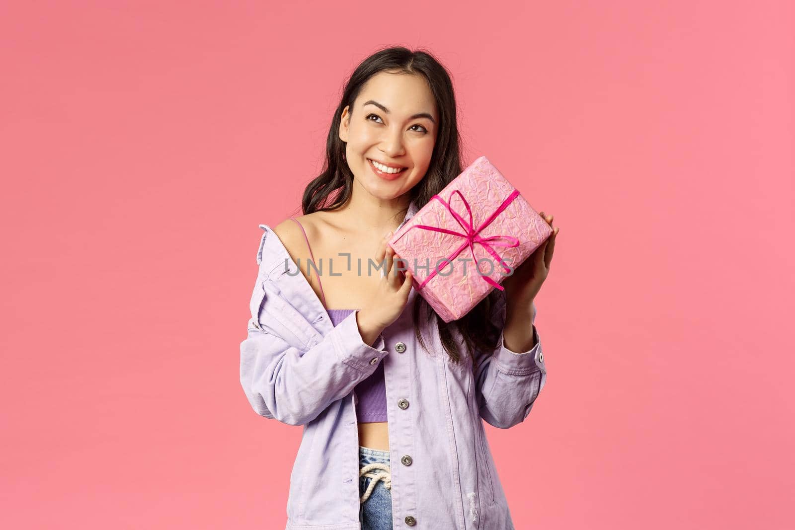 Curious whats inside. Portrait of cute silly asian girl shaking box with gift to guess what is it, smiling amused, leaning to present with ear trying hear movement, standing pink background by Benzoix