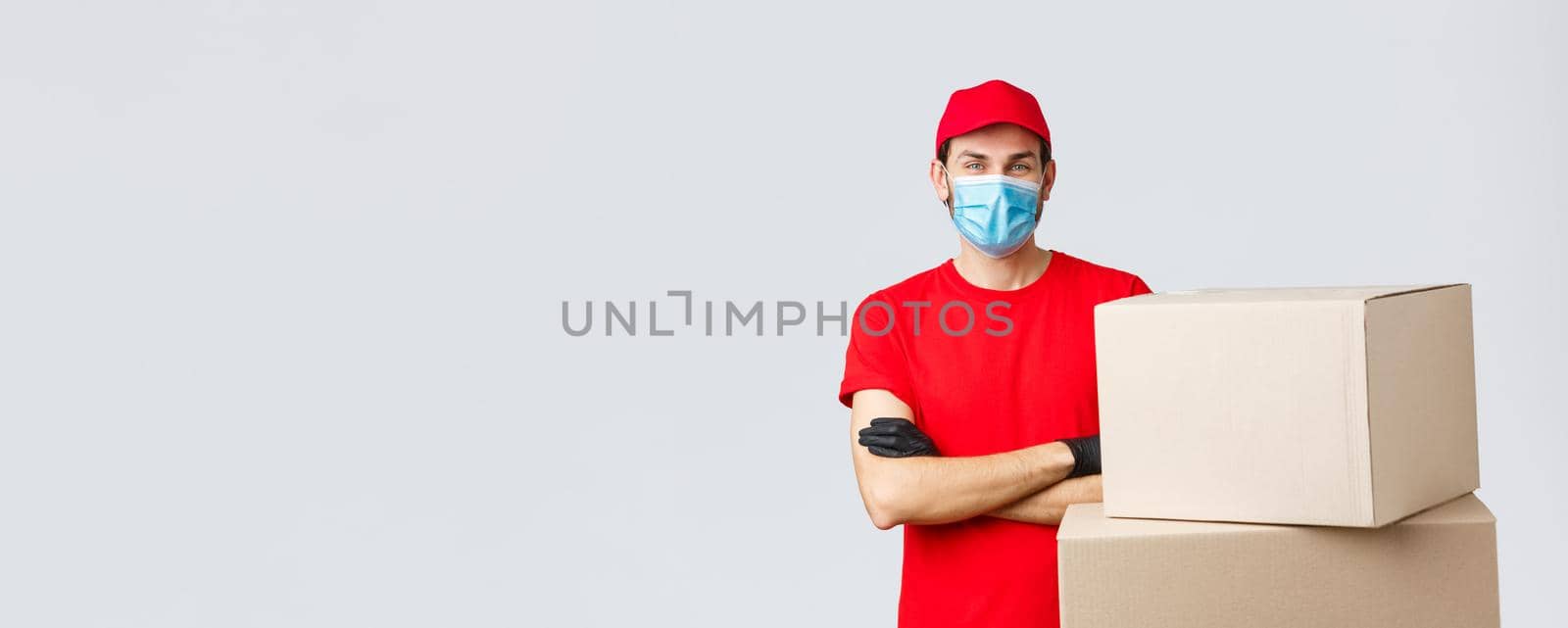 Packages and parcels delivery, covid-19 quarantine and transfer orders. Confident young courier in red uniform, gloves and medical mask, cross arms as standing boxes, grey background.