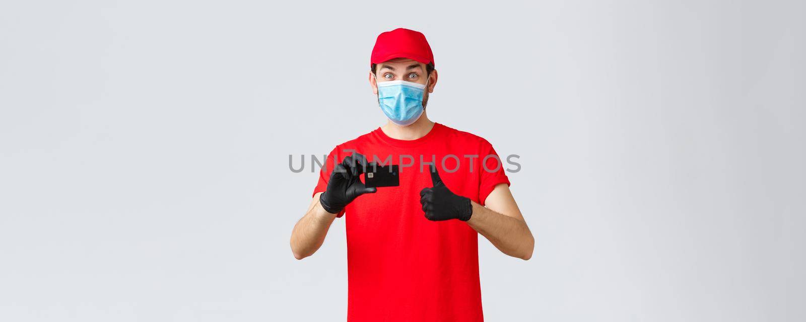 Contactless delivery, payment and online shopping during covid-19, self-quarantine. Excited happy courier in red t-shirt and cap uniform, face mask with gloves, recommend credit card order paying.