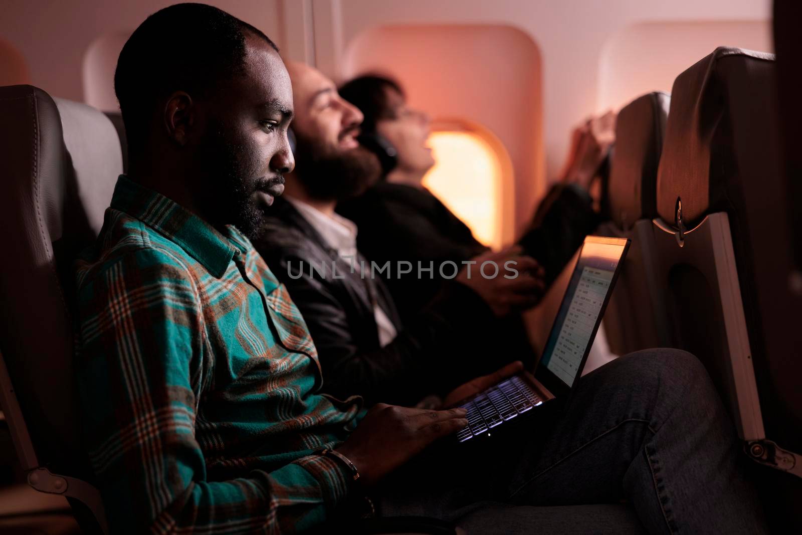 African american man travelling abroad by airplane on international flight, using airline service and aerial transportation. Freelancer using laptop computer during sunset in economy class.