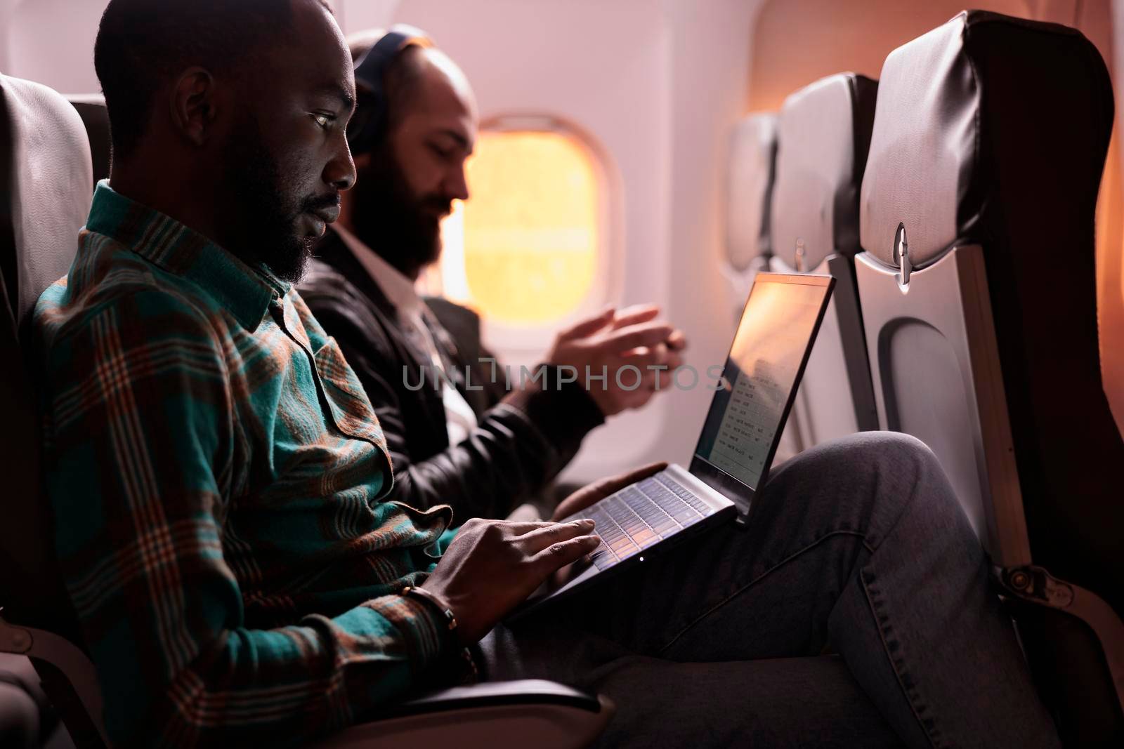 African american male freelancer working on laptop during sunset flight, waiting to arrive at holiday destination. Flying in economy class with group of tourists, using computer online.