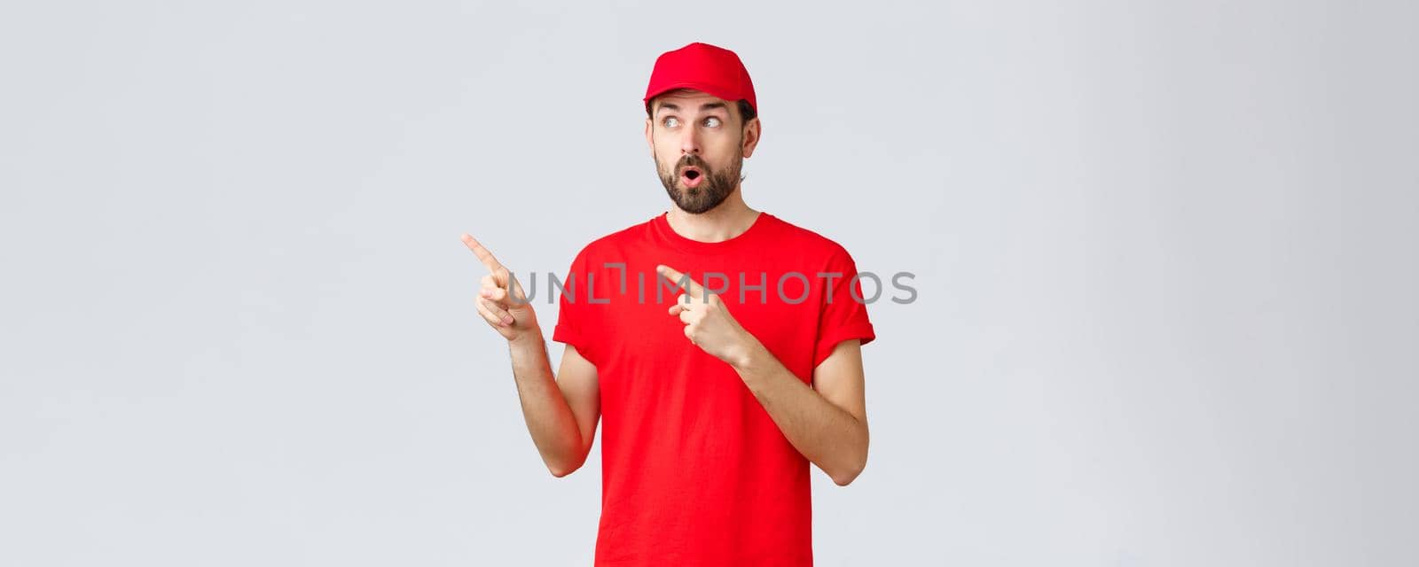 Online shopping, delivery during quarantine and takeaway concept. Impressed and wondered courier in red t-shirt with cap, employee uniform, open mouth shocked and staring left, pointing fingers by Benzoix