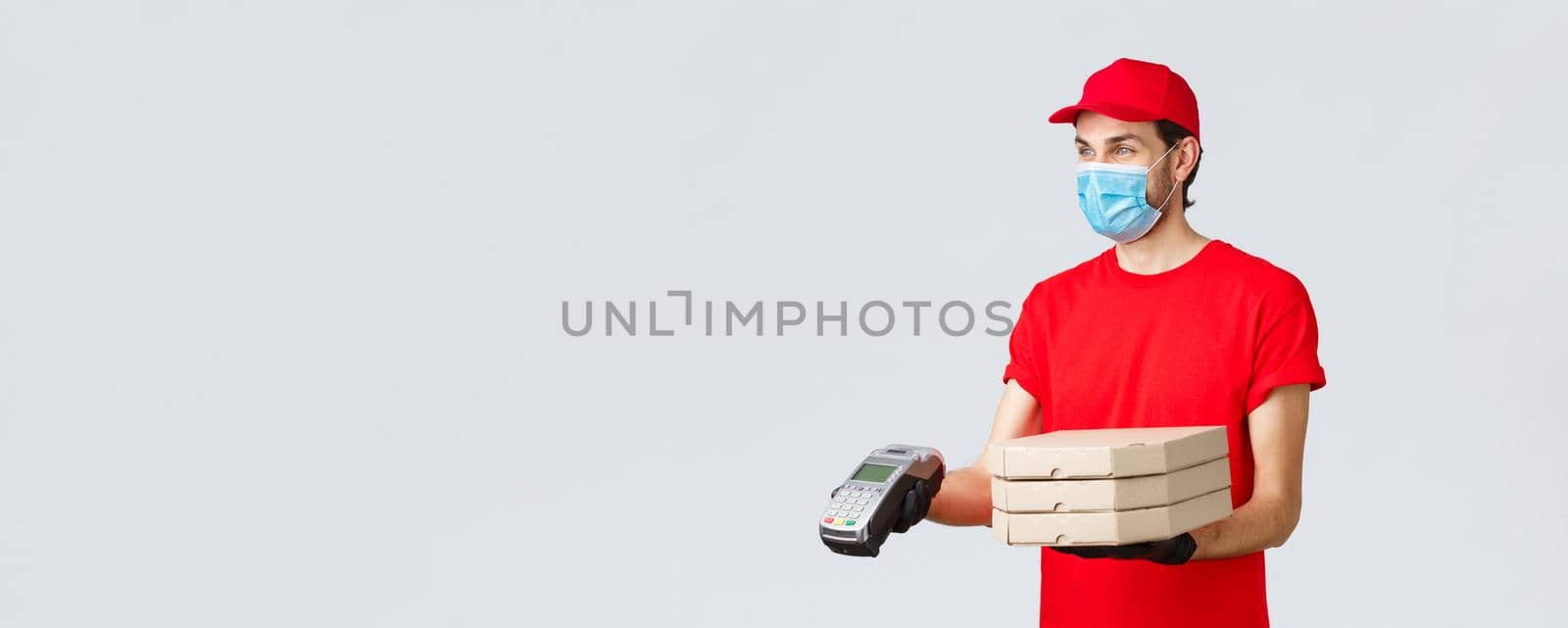 Food delivery, application, online grocery, contactless shopping and covid-19 concept. Pleasant delivery guy in red uniform, gloves and face mask, giving order client with POS terminal, deliver pizza.