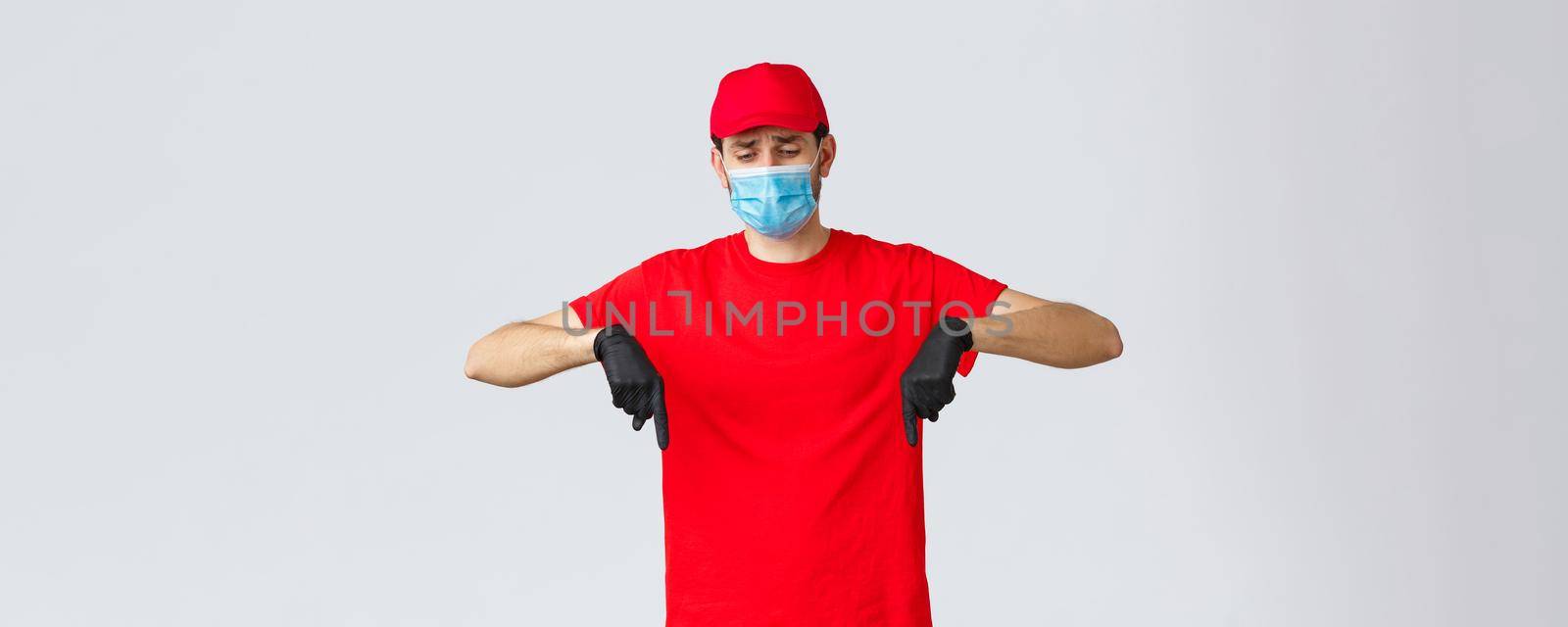 Covid-19, self-quarantine, online shopping and shipping concept. Upset gloomy courier feeling sad looking down and pointing bottom advertisement, frowning, wear face mask and gloves.