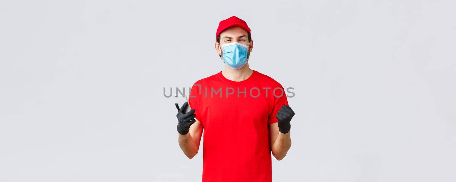 Contactless delivery, payment and online shopping during covid-19, self-quarantine. Rejoicing, happy courier in red uniform, gloves and mask, smiling, read good news in application, mobile phone.