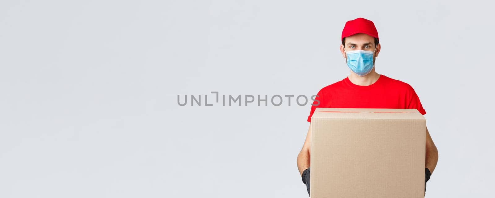 Groceries and packages delivery, covid-19, quarantine and shopping concept. Serious courier in red uniform, gloves and protective face mask, deliver package box to client house during coronavirus by Benzoix