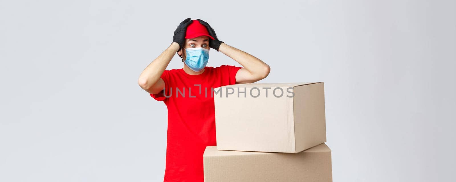 Packages and parcels delivery, covid-19 quarantine and transfer orders. Concerned and troubled courier in red uniform, face mask and gloves, grab head and gasping shocked staring at boxes.