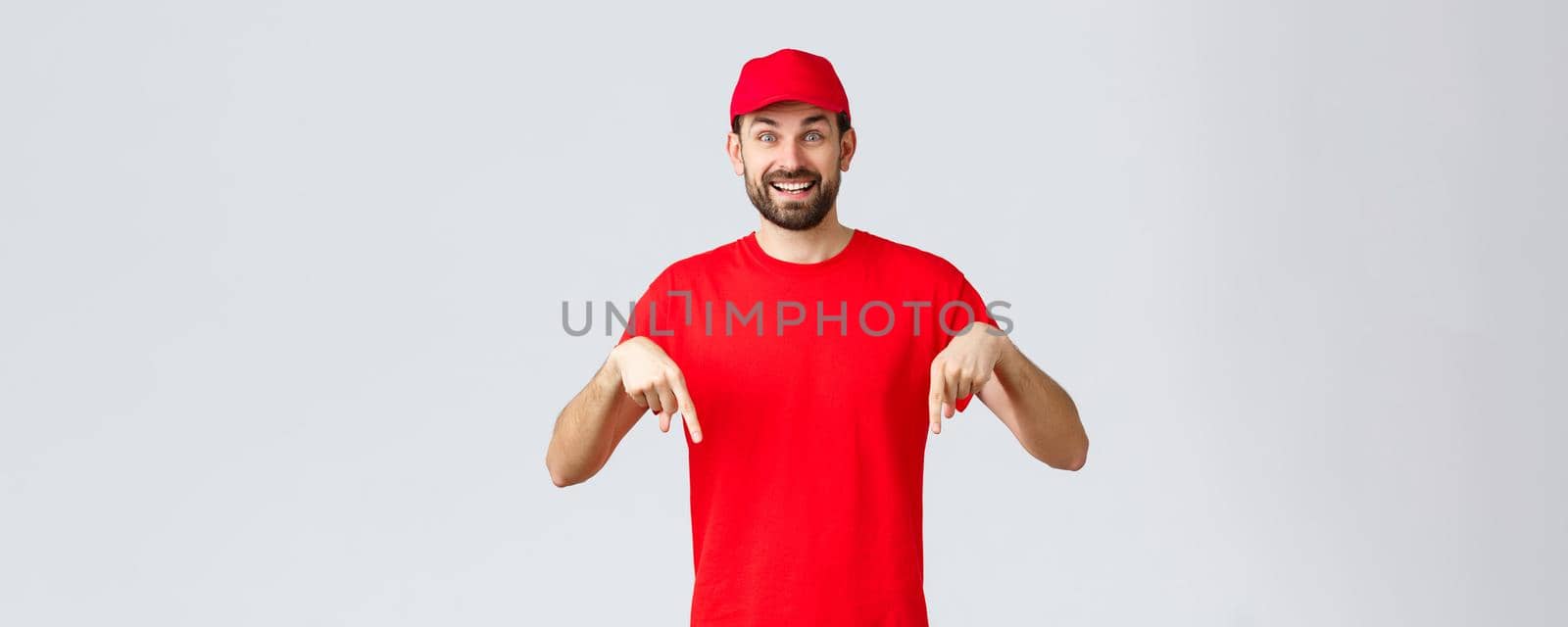 Online shopping, delivery during quarantine and takeaway concept. Cheerful smiling, amused courier in red uniform cap and t-shirt, excited about new promo, pointing fingers down, grey background.