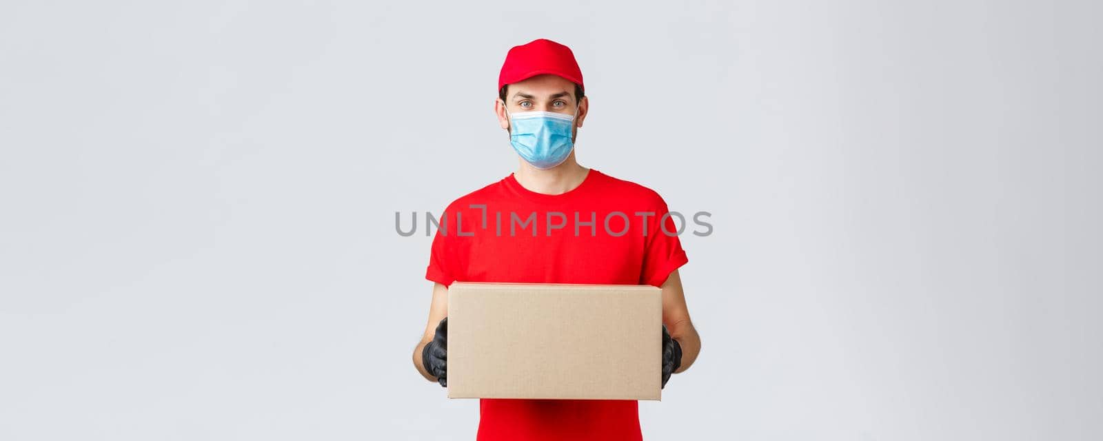 Packages and parcels delivery, covid-19 self-quarantine delivery, transfer orders. Young courier in red uniform, gloves and face mask, holding box, give-out order to client, contactless service.