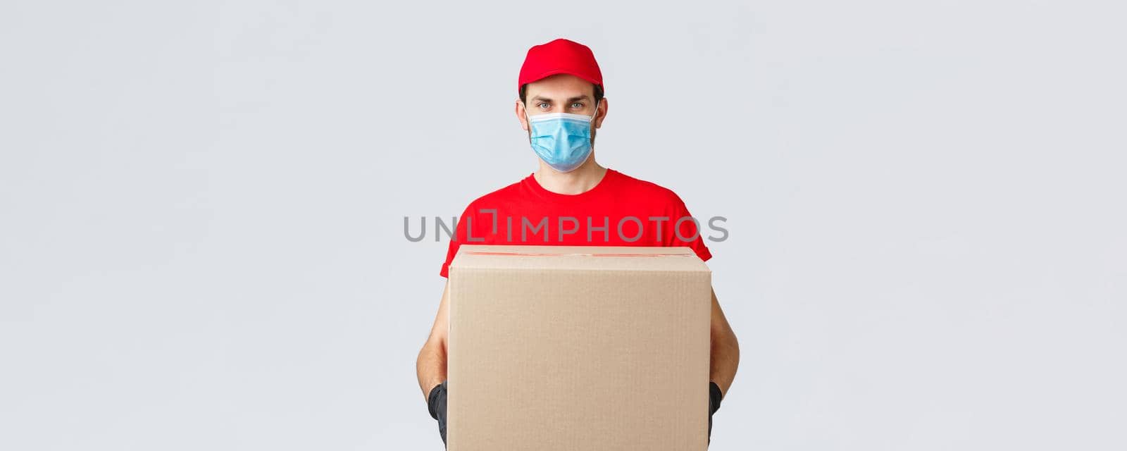 Groceries and packages delivery, covid-19, quarantine and shopping concept. Serious courier in red uniform, gloves and protective face mask, deliver package box to client house during coronavirus.