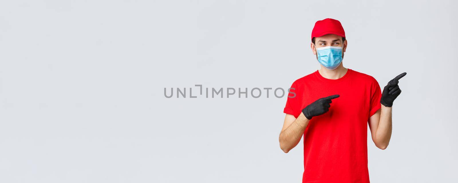 Covid-19, self-quarantine, online shopping and shipping concept. Intrigued smiling delivery guy in red t-shirt, cap carrier uniform, using medical mask and gloves to delivery to client, point right by Benzoix