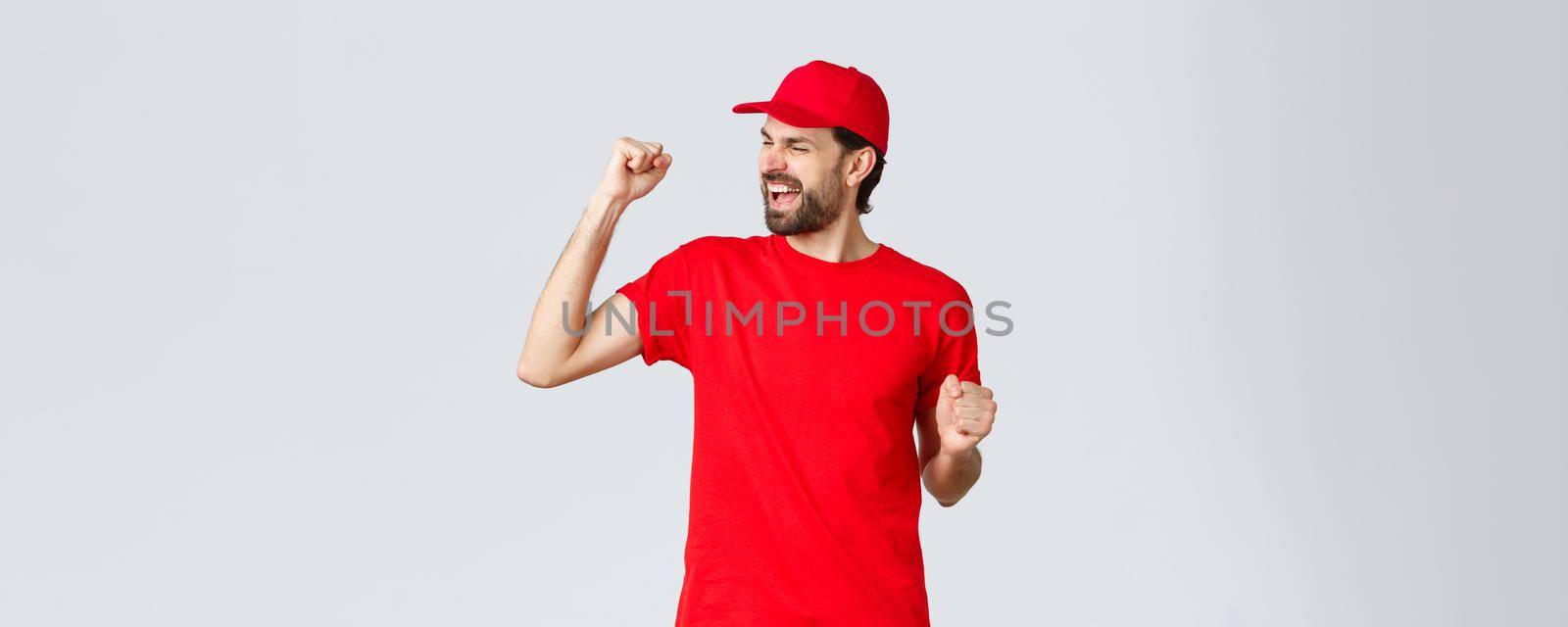 Cheerful and carefree young bearded delivery guy in red uniform cap and t-shirt, singing, yelling yes and fist pump in celebration, triumphing and rejoicing over great news, grey background.