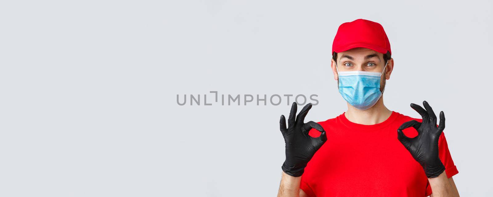 Contactless delivering, covid-19 and shopping concept. Cheerful, pleased courier in red uniform, cap and medical mask with gloves show okay, approval or guarantee gesture, recommend service by Benzoix