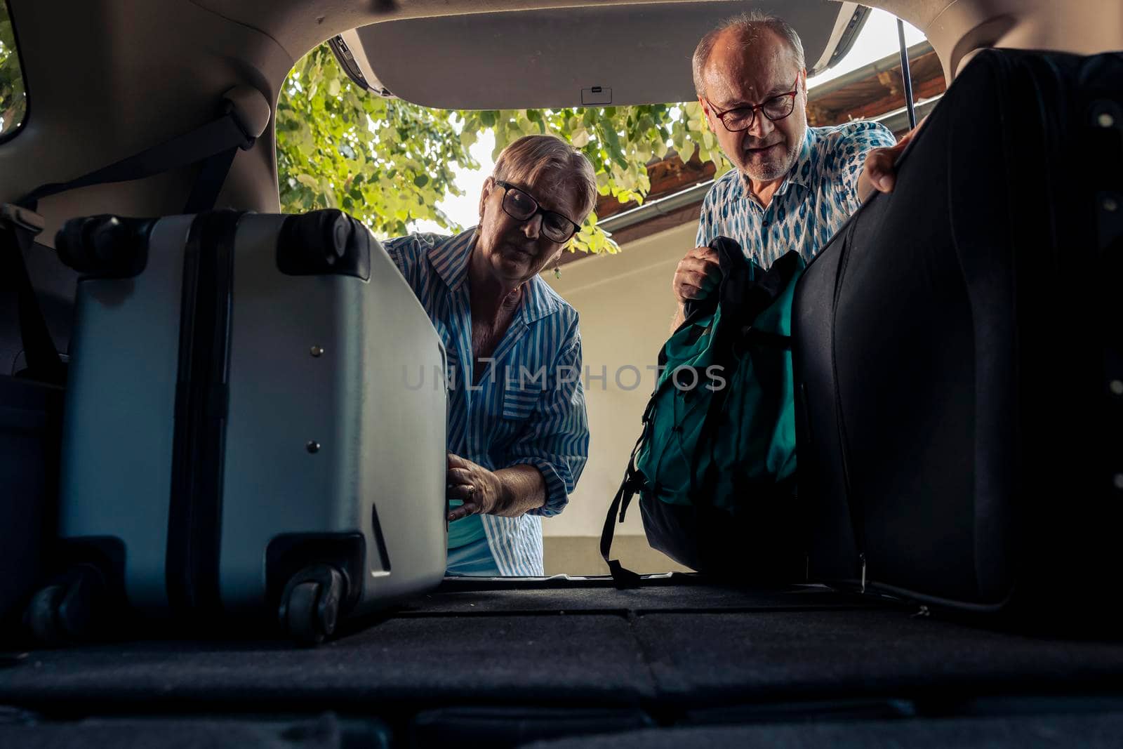 Senior couple loading bags in car trunk, preparing to leave on summer holiday with vehicle. People travelling on vacation road trip in retirement age, going on adventure destination.