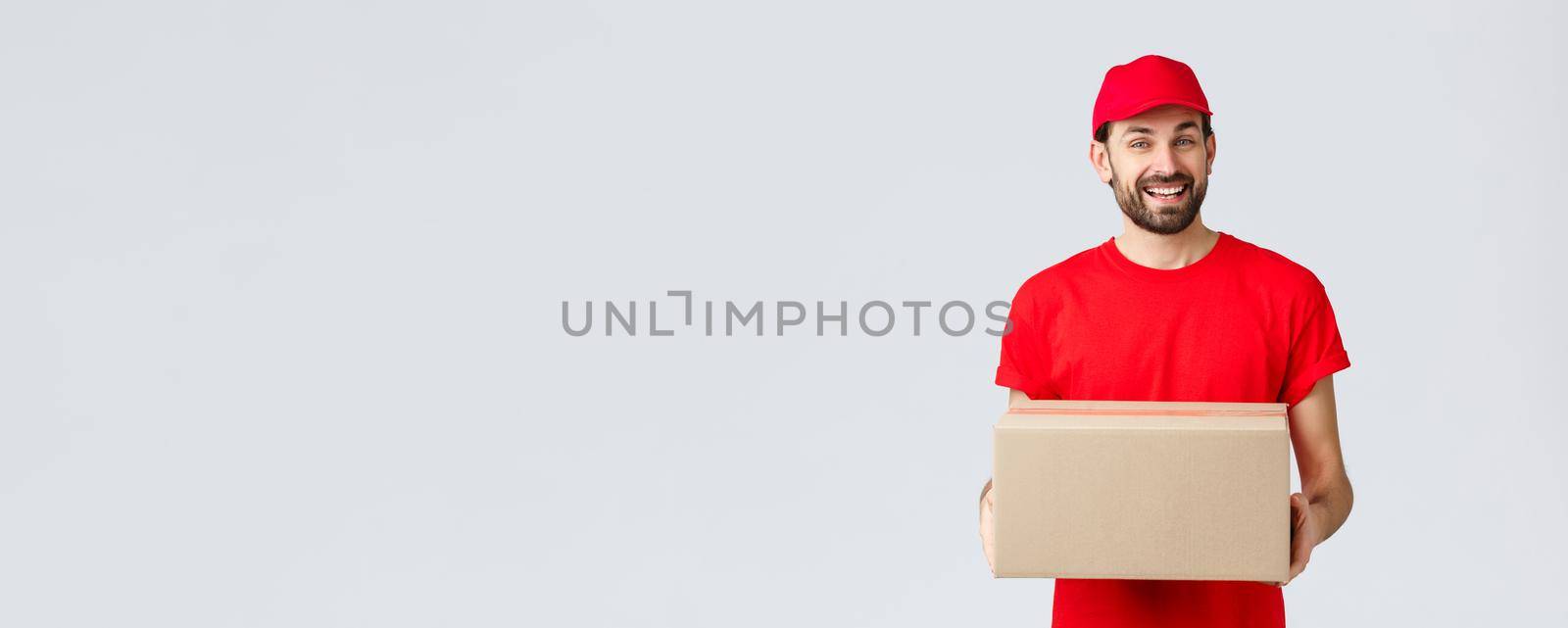 Order delivery, online shopping and package shipping concept. Friendly smiling courier in red uniform cap and t-shirt, handing out packages for customers. Employee bring parcel box, grey background.