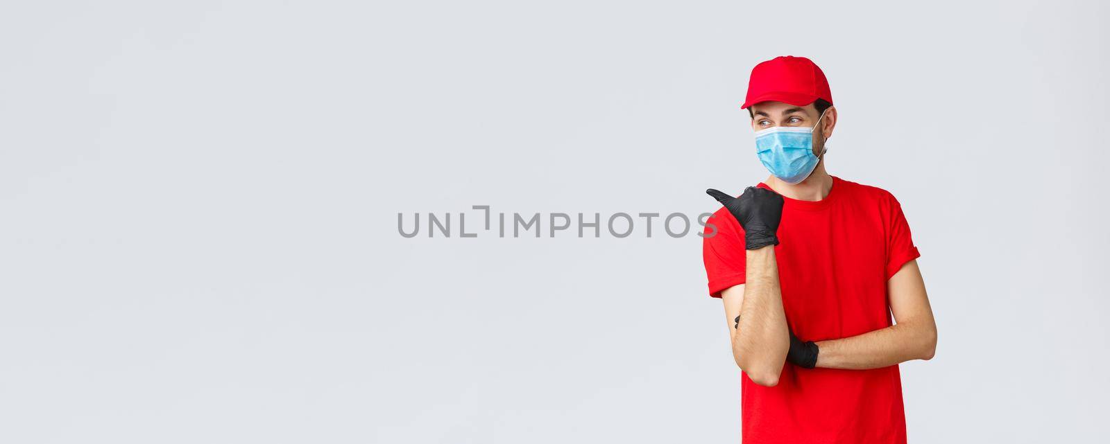 Covid-19, self-quarantine, online shopping and shipping concept. Delivery guy in red uniform, gloves and face mask, pointing left and smiling, recommend client use courier service.