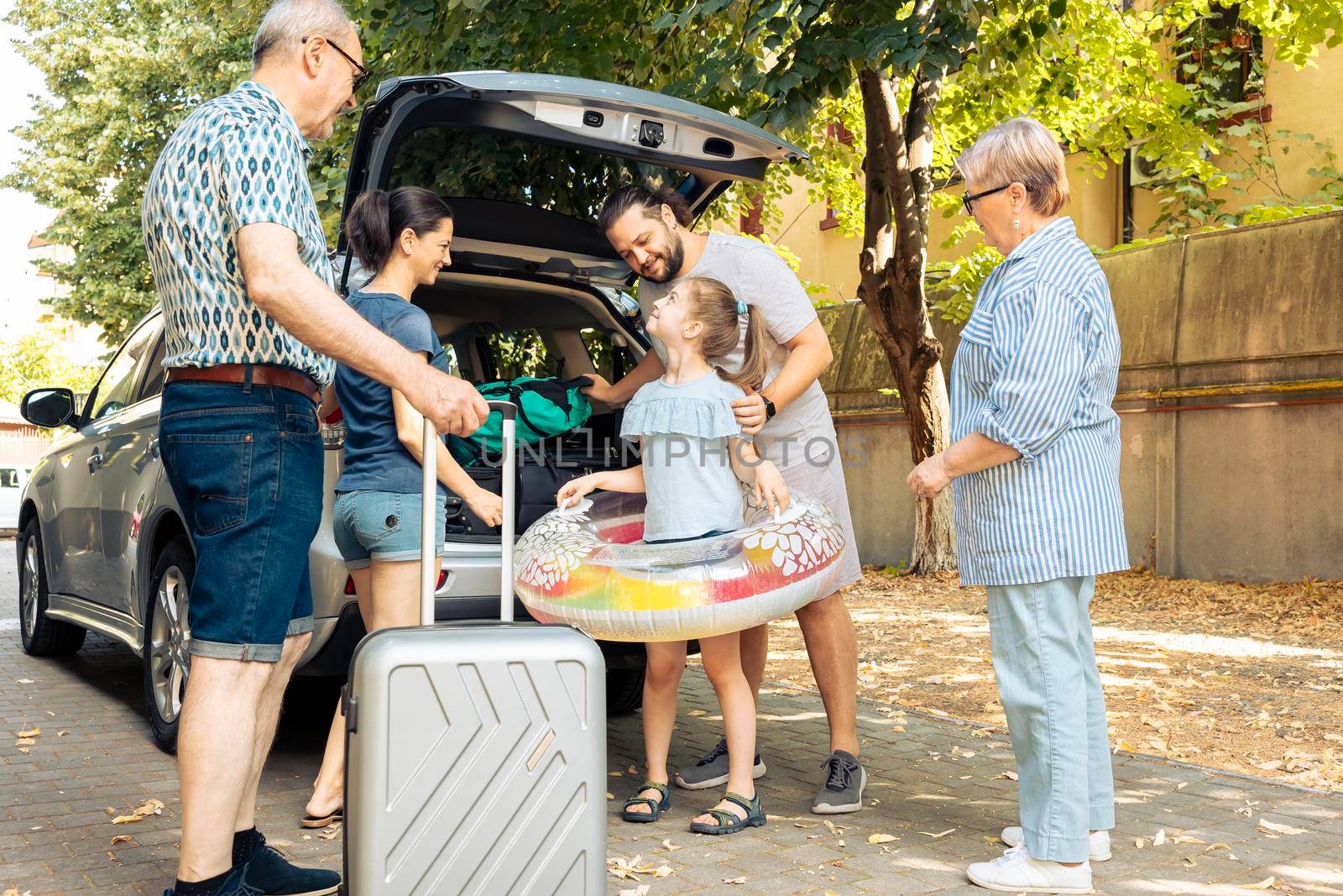 Happy family travelling at seaside with car, leaving on vacation trip with parents, grandparents and small girl. Loading luggage and suitcase in automobile trunk, summer adventure.