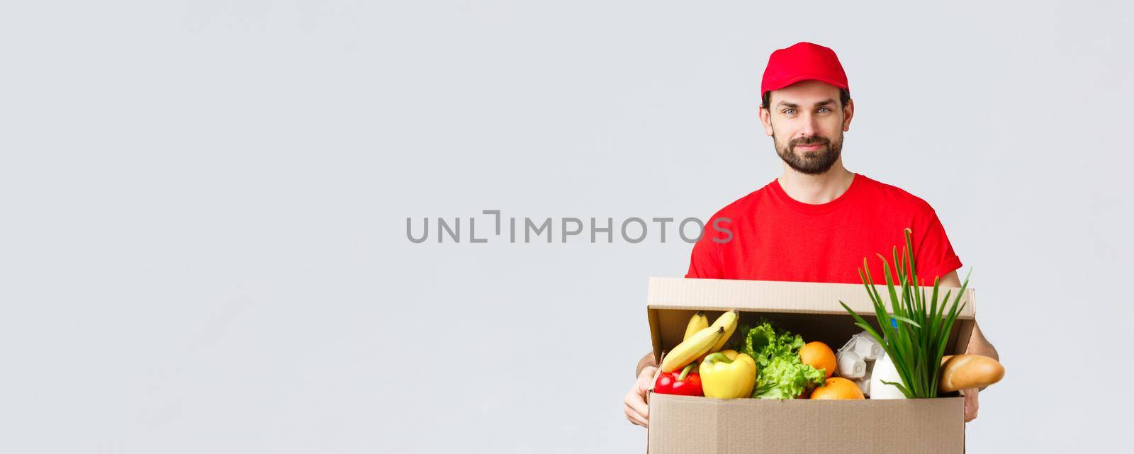 Groceries and packages delivery, covid-19, quarantine and shopping concept. Handsome courier in red uniform cap and t-shirt, bring grocery package, box delivery to client house, fast shipping.