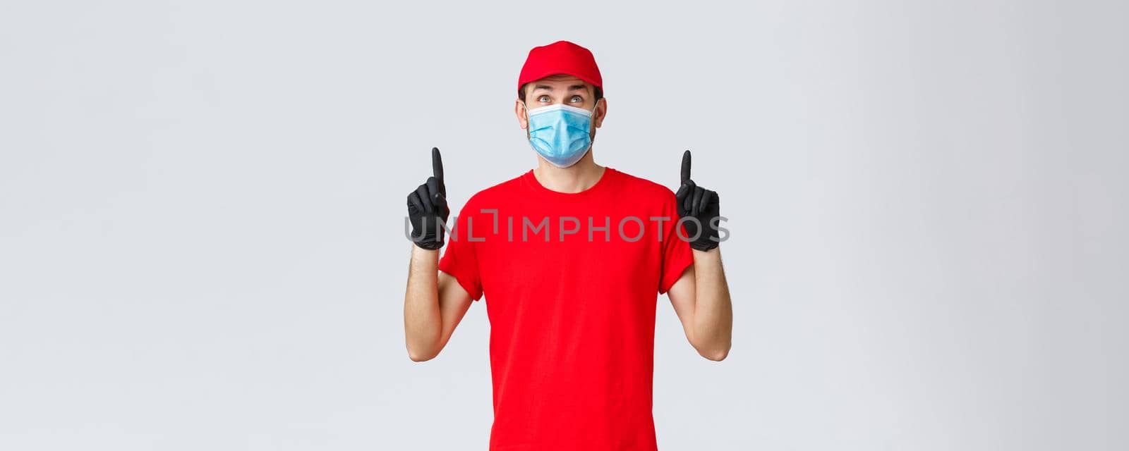 Covid-19, self-quarantine, online shopping and shipping concept. Smiling delivery guy red uniform cap and t-shirt, medical mask with rubber gloves, looking pointing up curious, reading sign or promo by Benzoix