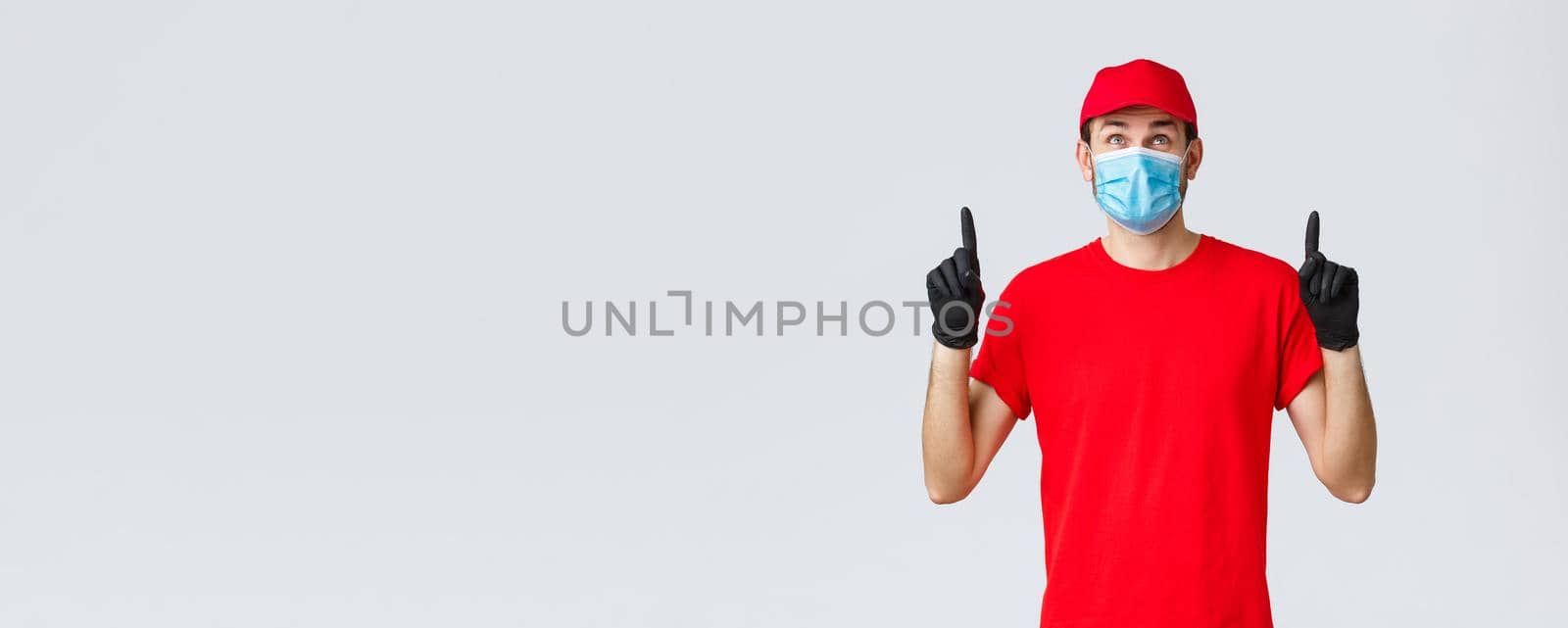 Covid-19, self-quarantine, online shopping and shipping concept. Smiling delivery guy red uniform cap and t-shirt, medical mask with rubber gloves, looking pointing up curious, reading sign or promo by Benzoix