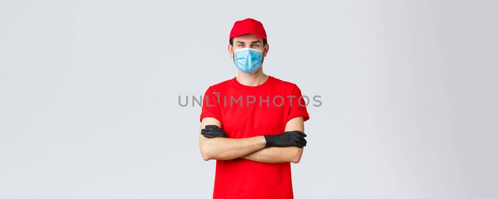 Covid-19, self-quarantine, online shopping and shipping concept. Confident smiling delivery man in red cap, t-shirt, wearing protective medical mask and rubber gloves while making courier order.