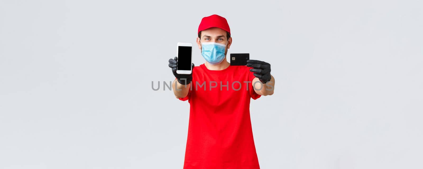 Contactless delivery, payment and online shopping during covid-19, self-quarantine. Friendly courier in red uniform, face mask and gloves, showing smartphone screen and credit card, order internet.