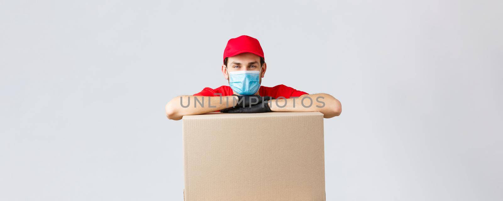 Packages and parcels delivery, covid-19 quarantine and transfer orders. Young courier in red uniform cap, face mask and gloves, leaning on box to deliver, shipping your orders, grey background.