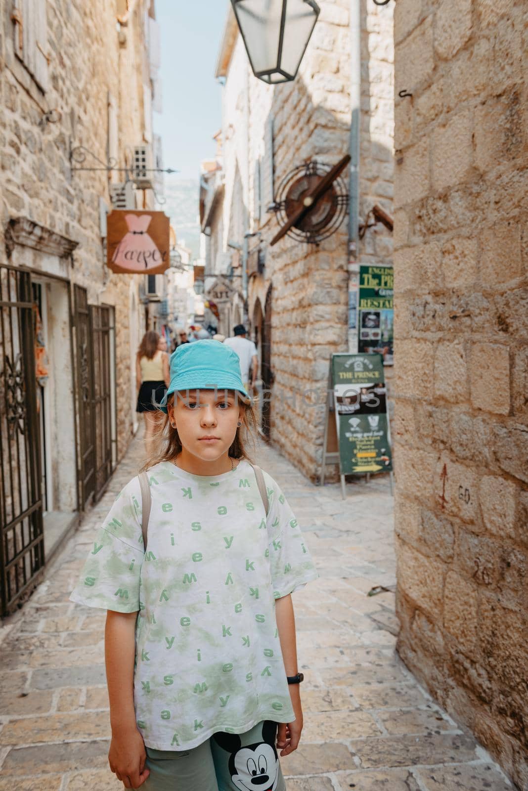 Girl Tourist Walking Through Ancient Narrow Street On A Beautiful Summer Day In Mediterranean Medieval City, Old Town Budva, Montenegro. Young Beautiful Cheerful Woman Walking On Old Street At Tropical Town. Pretty Girl Looking At You And Smiling by Andrii_Ko