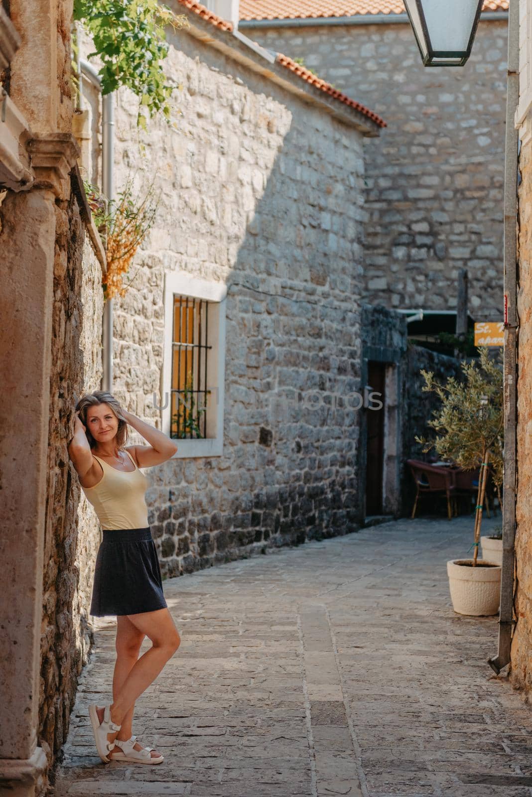 Girl Tourist Walking Through Ancient Narrow Street On A Beautiful Summer Day In MEDITERRANEAN MEDIEVAL CITY, OLD TOWN BUDVA, MONTENEGRO. Young Beautiful Cheerful Woman Walking On Old Street At Tropical Town. Pretty Girl Looking At You And Smiling by Andrii_Ko