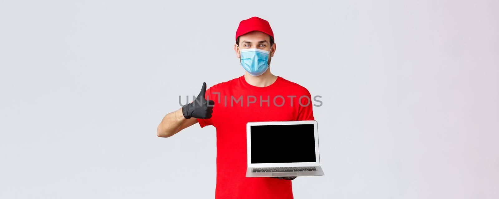 Customer support, covid-19 delivery packages, online orders processing concept. Cheerful courier in red uniform, medical face mask and gloves recommend webpage, show laptop screen and thumb-up.