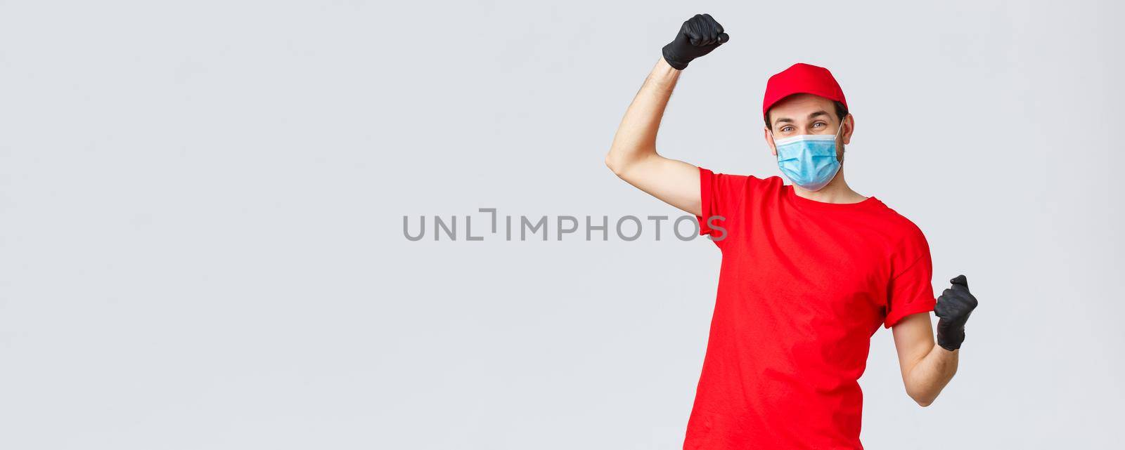 Groceries and packages delivery, covid-19, quarantine and shopping concept. Cheerful courier red uniform, gloves and face mask, raising hands up, chanting rejoicing over good news, fight coronavirus.