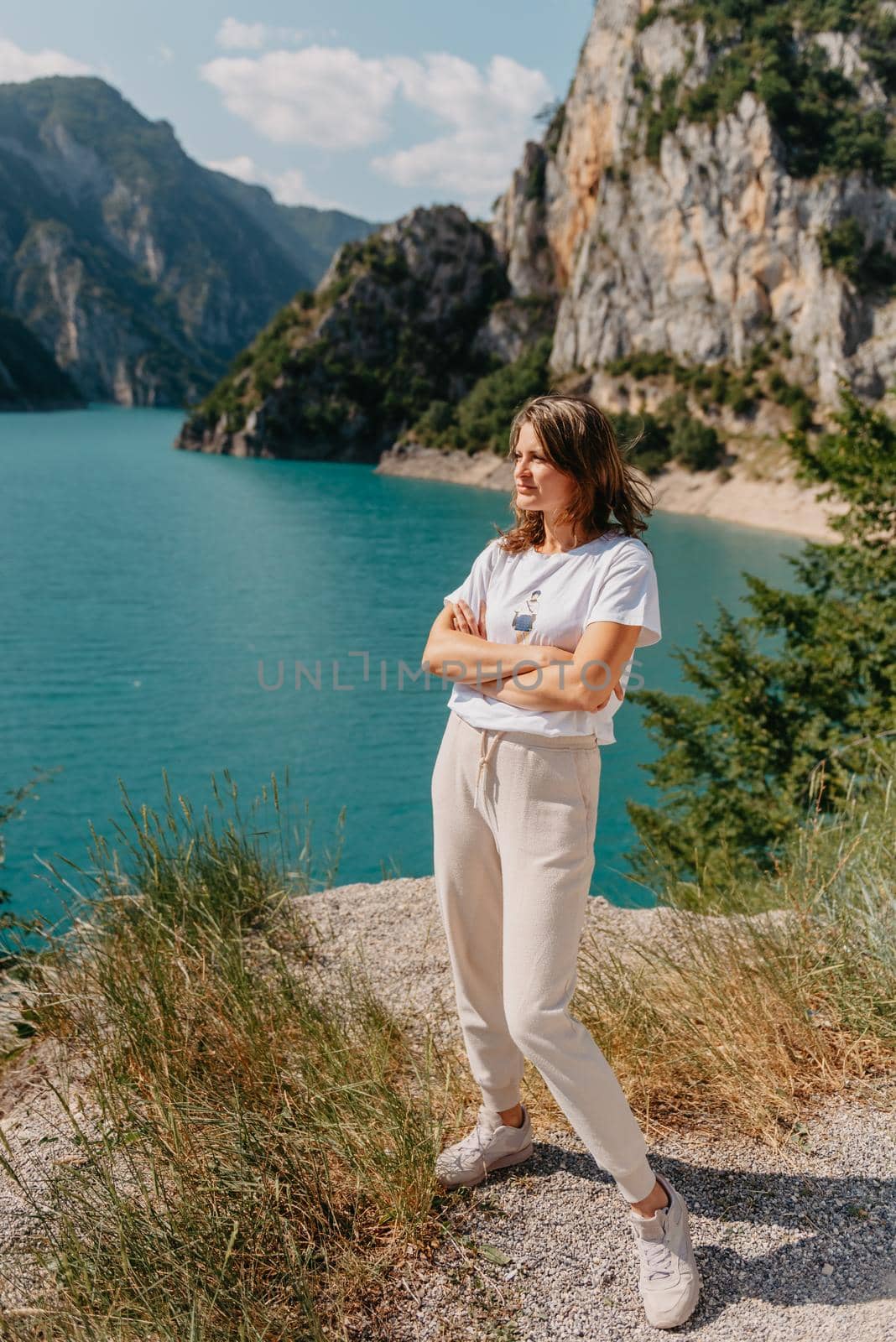 Girl tourist stands on the shore of a lake in the mountains. beautiful landscape, Piva Lake in Montenegro.. Woman is standing on the coast of Piva lake at sunset in summer. Landscape with girl, famous lake with beautiful reflection in water, trees, sky with clouds. Travel by Andrii_Ko