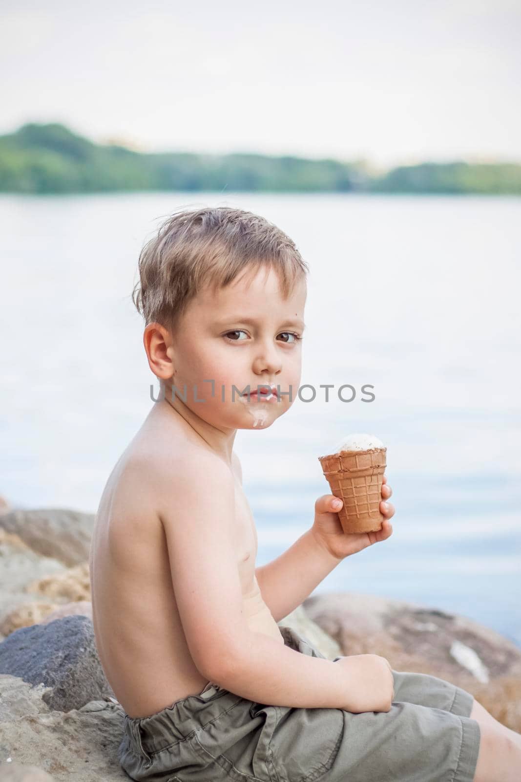 A cute blond boy appetizingly eats ice cream in the summer, sitting on the bank of the river. Cool off by the water. Funny facial expression. summer heat