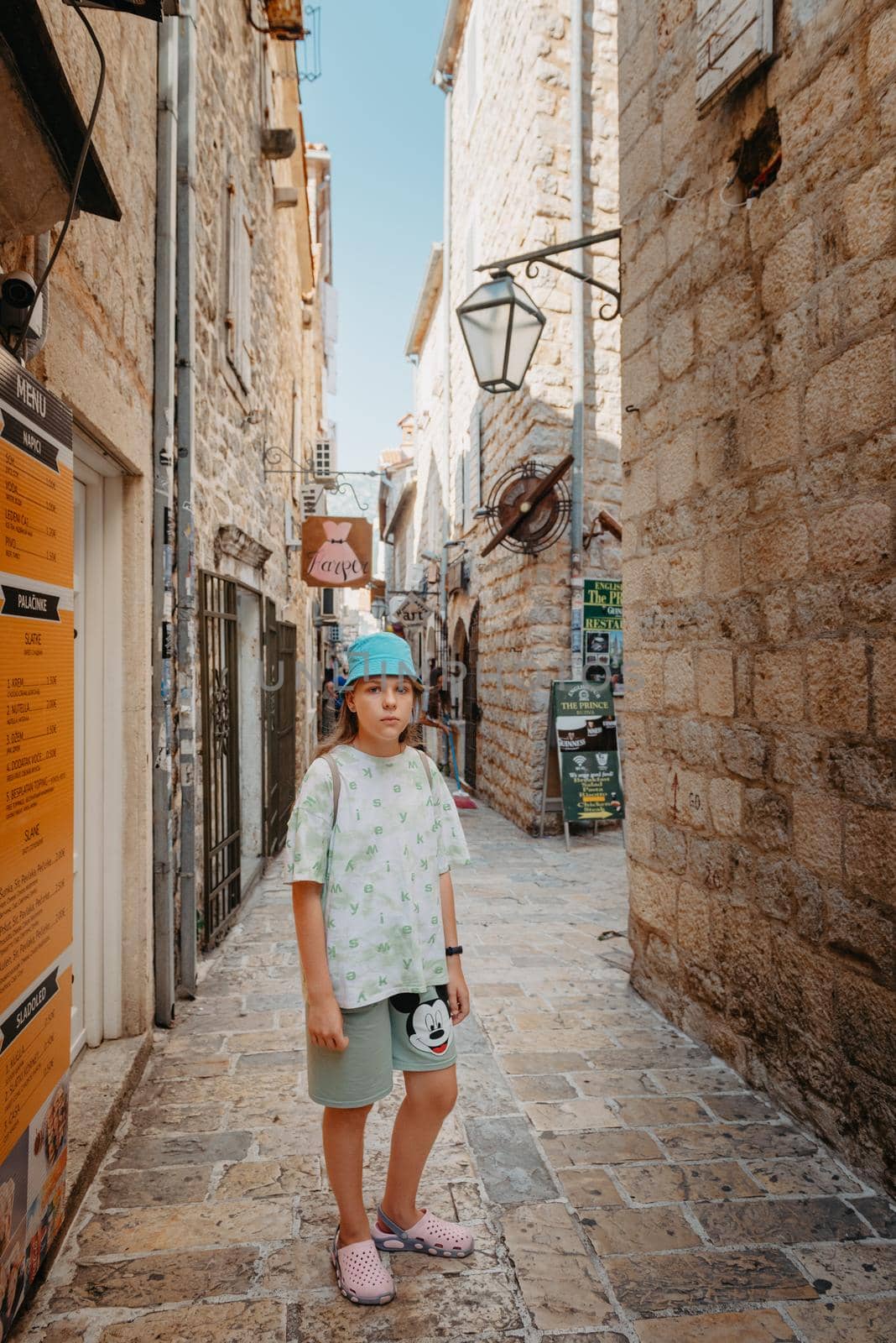 Girl tourist walking through ancient narrow street on a beautiful summer day in MEDITERRANEAN MEDIEVAL CITY , OLD TOWN bUDVA, MONTENEGRO. Young beautiful cheerful woman walking on old street at tropical town. Pretty girl looking at you and smiling