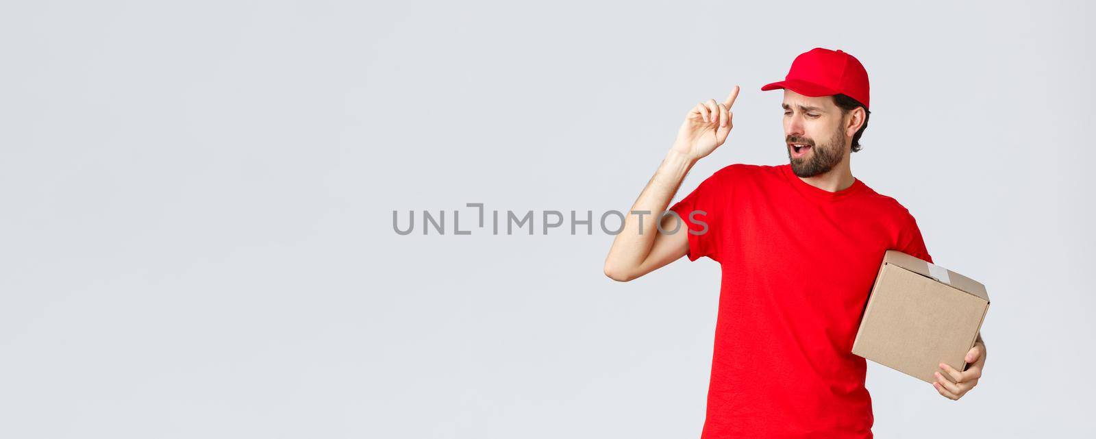 Order delivery, online shopping and package shipping concept. Carefree handsome bearded courier in red uniform, cap and t-shirt, holding box package, singing song, grey background.